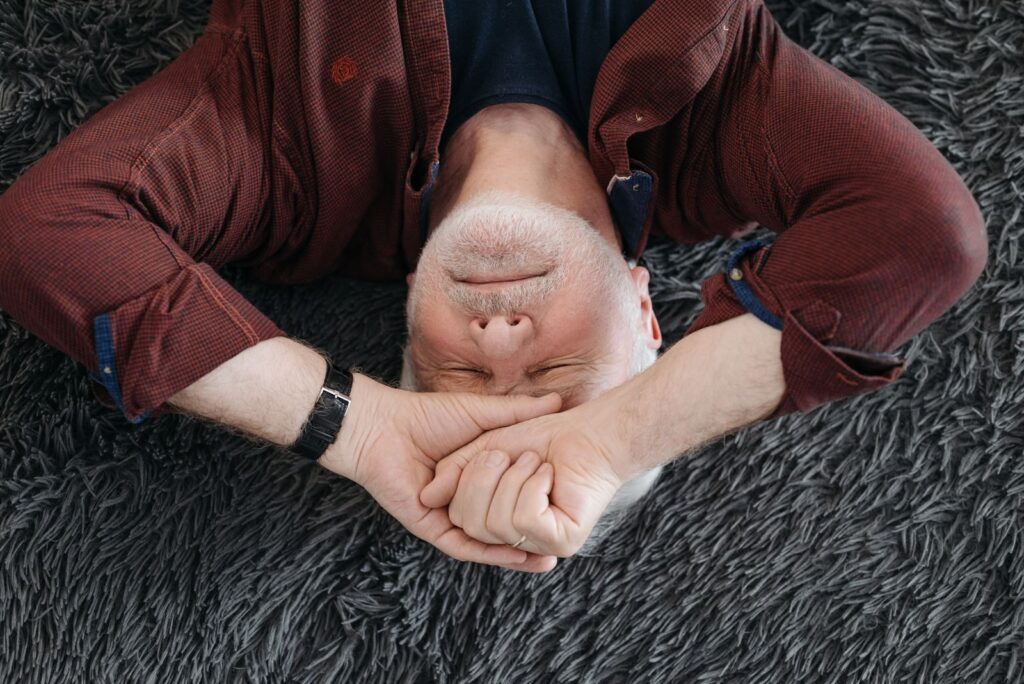 A senior man lying on a soft surface indoors with closed eyes, expressing a range of emotions.