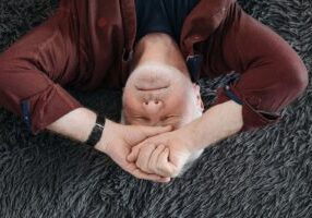 A senior man lying on a soft surface indoors with closed eyes, expressing a range of emotions.